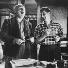 Zwei Männer lachen, umgeben von einem Schreibtisch mit Papieren und einem Bücherregal. Im Hintergrund ist eine Landkarte.