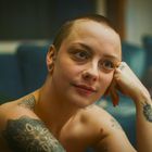 Eine Person mit kurzen Haaren, Tattoos und freundlichem Gesichtsausdruck stützt den Kopf auf ihre Hand. Hintergrund unscharf.