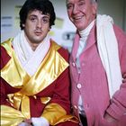 Ein Mann in einem roten und goldenen Boxermantel sitzt neben einem älteren Mann in einer rosa Strickjacke, beide lächeln.