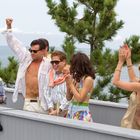Sechs Menschen feiern auf einer Terrasse am Meer; einige heben die Arme, tragen Sonnenbrillen, im Hintergrund Bäume.