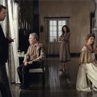 Vier Personen in einem Raum; ein Mann steht, ein anderer sitzt, zwei Frauen sitzen und stehen, alle blicken ernst.