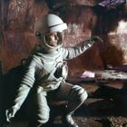 „DEFA“-Logo in der Ecke. Ein Astronaut in einem Raumanzug kauert in einer futuristischen, rostigen Umgebung.