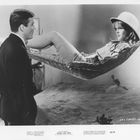 BRIGITTE BARDOT in PLEASE, NOT NOW. Ein Mann steht vor einer Frau, die entspannt in einer Hängematte liegt.