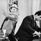 „FBI Agent Eddie wieder Colt-richtig“ steht unten. Zwei elegant gekleidete Männer, einer mit Fedora, lachen in einem Raum.