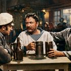 Drei Männer in Seemannskleidung sitzen an einem Tisch mit Bierkrügen in einer rustikalen Taverne. Sie wirken vertieft im Gespräch.