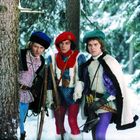 Drei Männer in bunten mittelalterlichen Kostümen im verschneiten Wald. Sie halten Ski und tragen Mützen.
