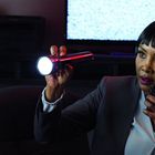 Frau in Anzug mit "Edwina 'Clue' Cummings" Namensschild, hält Taschenlampe und Funkgerät, beobachtet Monitorrauschen.