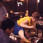 Eine Frau in gelbem Top tritt mit ihrem Bein einen Tisch um, es sind Mahjong-Steine sichtbar. Menschen stehen im Hintergrund.