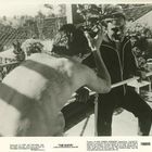 Zwei Männer unterhalten sich draußen; einer ist unbekleidet, der andere trägt einen Trainingsanzug. Viel Sonnenlicht.