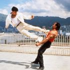 Zwei Männer kämpfen mit einem fliegenden Tritt auf einer Terrasse vor städtischer Skyline und Bergen.