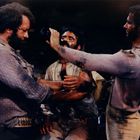 Drei Männer in dreckigen, abgetragenen Kleidern; einer gießt einem anderen etwas aus einer Flasche ein.