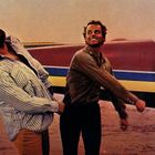 Zwei Männer, einer in gestreiftem Hemd, der andere in dunklem Hemd, stehen vor einem kleinen Flugzeug.