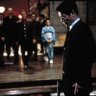 Ein Mann im Anzug steht im Vordergrund, mehrere Personen treten im Hintergrund aus einem rot beleuchteten Bereich auf.