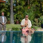 Ein Mann sitzt am Pool, trägt Sonnenbrille und Handtuch, daneben steht ein Mann im Anzug. Hintergrund: Palmen und Pflanzen.