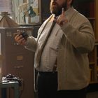 Ein Mann mit Bart und Brille in beiger Strickjacke spricht in einem Büro und hält ein kleines Objekt in der Hand.