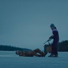 Ein Mensch liegt auf einem zugefrorenen See, während eine andere Person daneben steht, umgeben von einer bewaldeten Landschaft.