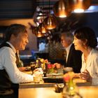 Zwei Menschen sitzen an einem Tisch in einem stilvollen Restaurant, umgeben von warmem Licht und Kupferlampen.