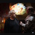 Zwei Männer stehen in einem bunten Raum, einer in schwarzem Shirt, der andere in einer glänzenden Rüstung.