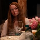 Eine nachdenkliche Frau mit langen roten Haaren sitzt an einem gedeckten Tisch mit einem Strauß rosa Tulpen im Vordergrund.