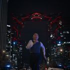 Eine Person rennt nachts in der Stadt. Oben leuchtet ein futuristisches, rot-illuminatiertes Objekt zwischen Hochhäusern.