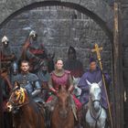 Ritter in Rüstungen reiten durch ein steinernes Tor; ein Priester hält ein Kreuz. Rustikale, mittelalterliche Atmosphäre.