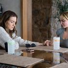 Zwei Frauen sitzen an einem Tisch und halten Hände. Vor ihnen sind Getränke und eine Medikamentenflasche. Sie wirken nachdenklich.