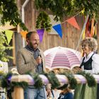 Zwei lachende Menschen auf einem Fest, umgeben von bunter Wimpel und einem dekorierten Maibaum. Eine Frau trägt Tracht.