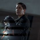 Ein Mann hält einen kleinen, zotteligen Hund in den Armen und eine Taschenlampe. Die Szene ist bei Regen und wirkt angespannt.