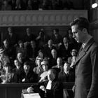 Ein Gerichtssaal mit einem Mann im Anzug im Vordergrund, Publikum im Hintergrund beobachtet aufmerksam. Schwarz-weiß.