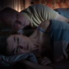 Ein älteres Paar liegt nebeneinander im Bett, in gedämpftem Licht, nachdenklich und eng umschlungen.