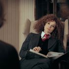 Eine Frau mit lockigem Haar sitzt nachdenklich in einem Sessel, hält einen geöffneten Aktenordner.