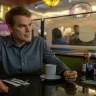 Eine Person in einem Diner, hält einen Becher. Im Hintergrund ist ein Burger-Symbol auf einer Trennscheibe abgebildet.