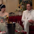 Eine Frau in einem floralen Kleid und ein Mann im weißen Anzug sitzen auf einem roten Sofa, mit Sekt und Blumen.