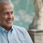 Ich sehe keine Texte. Ein älterer Mann mit grauen Haaren lächelt. Er trägt ein hellblaues Hemd. Im Hintergrund Natur.