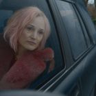 Über einem Rücken mit rosa Fellmantel lehnt eine Frau mit rosa Haaren am Autofenster, schaut nachdenklich hinaus.