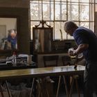 Ein Mann malt eine Holzplatte in einem Atelier. Spiegel, Gemälde und Arbeitsutensilien sind um ihn herum.