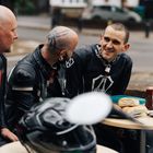 Vier Männer in Motorradkleidung sitzen an einem Tisch im Freien, mit Brötchen und Saucenflaschen darauf.