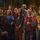 Eine Gruppe von Menschen singt zusammen in einem gemütlichen Raum mit antiken Uhren an der Wand.