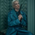 Ich kann keine spezifischen Texte oder Personen identifizieren. Eine Person in blauem Mantel hält einen Revolver und lehnt an dekorativer Wand.