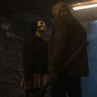 Eine Person mit blonden Haaren in blauem Mantel hält nachdenklich einen Revolver, vor dekorativer Wand.