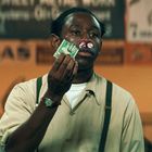 Ein Mann mit Wattestäbchen und blutverschmierten Nasenlöchern hält Geldscheine. Im Hintergrund unscharfe Schilder.