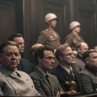 Ein Raum mit Männern in militärischen Uniformen und Anzügen, einige sitzen auf Bänken, andere stehen aufmerksam.