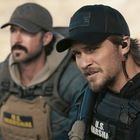 Mann mit schwarzer Kappe trägt Jacke mit "U.S. Marshal"-Aufschrift. Zweiter Mann unscharf im Hintergrund.