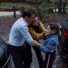 Drei Personen stehen nah beieinander, ein Mann, eine Frau und ein Mädchen, zwischen zwei Autos im Wald.