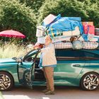 Ein Mann steht neben einem beladenen Auto, dessen Dach voll mit Gepäck ist; im Hintergrund ein roter Sonnenschirm.