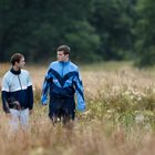 Zwei Männer in Sportjacken laufen durch ein hohes, wild wachsendes Feld mit Bäumen im Hintergrund.