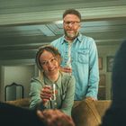 Zwei Personen sitzen auf einem Sofa mit Drinks. Eine Frau lächelt freundlich, ein Mann steht dahinter, ebenfalls mit einem Drink.