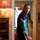 Ein Mann mit Schnurrbart steht in einem gemütlichen, leicht chaotischen Raum. Er trägt eine bunte Trainingsjacke.