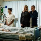 Im Krankenhauszimmer liegt eine Person im Bett, umgeben von medizinischem Gerät. Zwei Pflegerinnen und zwei Besucher stehen daneben.