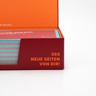 "365 NEUE SEITEN VON DIR!" steht auf einer roten Box mit sichtbaren bunten Papierstapeln darin.